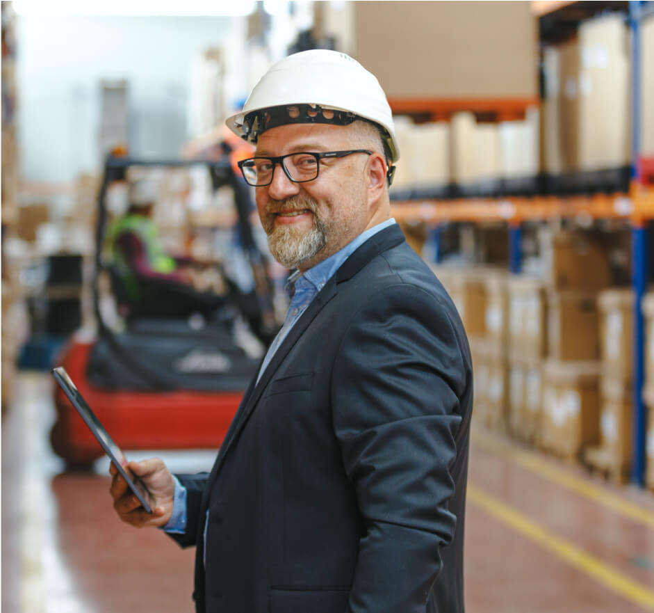 Middle aged man wearing a hard hat and a casual suit, smiling while holding a tablet inside a wear house.