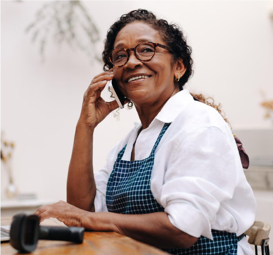 Middle aged woman sitting at a desk while talking on a cell phone wearing an apron.
